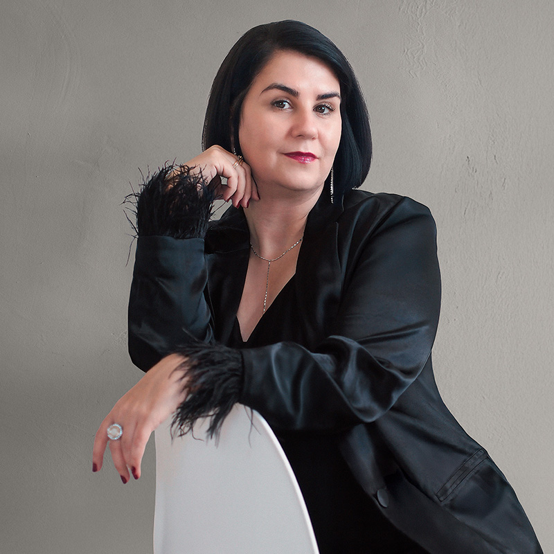 Karin Schön-von Hasseln, Stil- und Imageberaterin aus Stuttgart, sitzt selbstbewusst auf einem Stuhl und blickt ruhig in die Kamera. Sie trägt ein elegantes schwarzes Outfit mit Federbesatz an den Ärmeln.